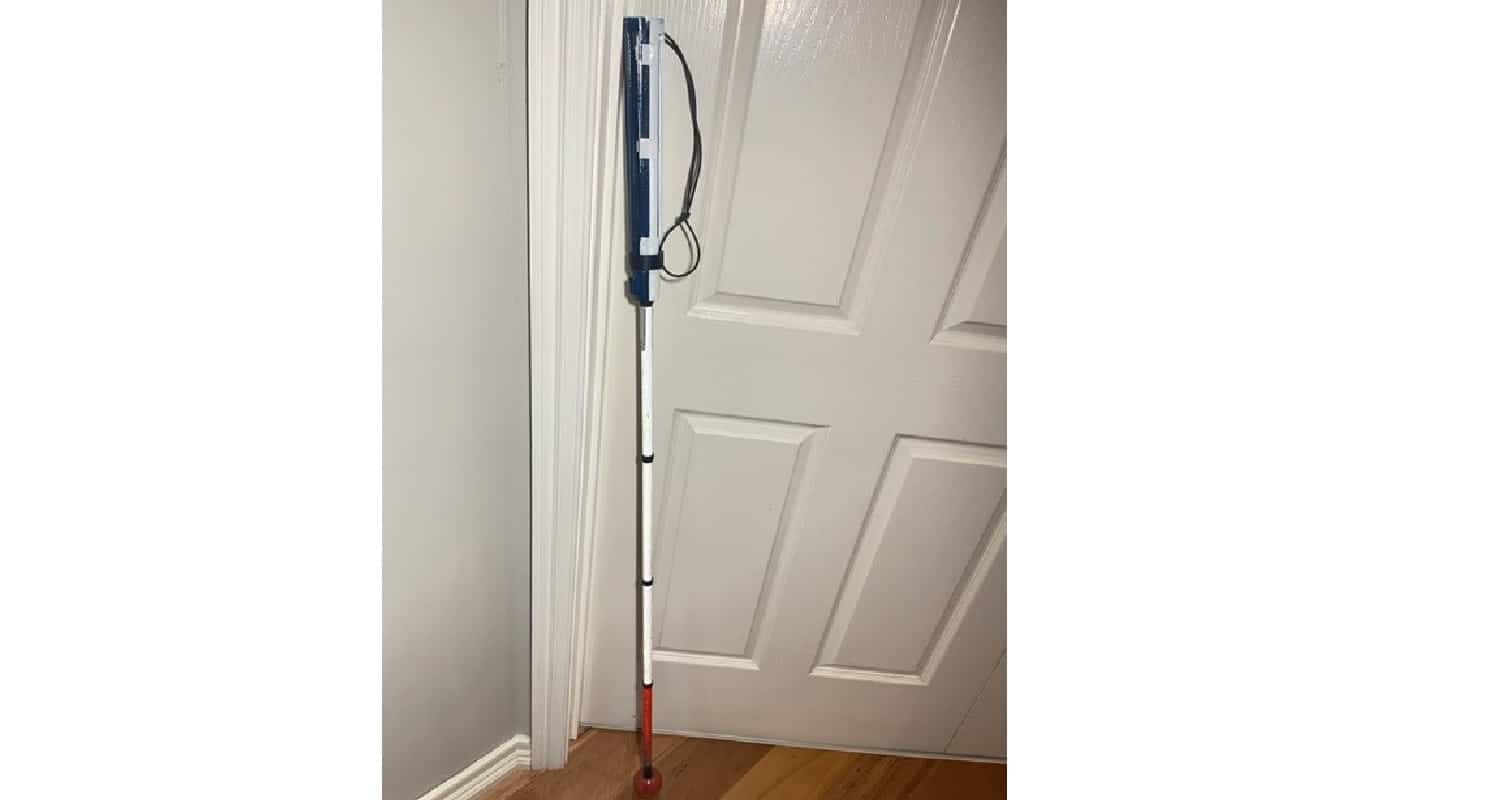 The image is a traditional white cane with a hand made model of the designed device clipped onto the handle of the cane.